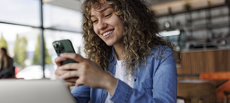 Young smiling woman using mobile phone while working on a laptop at a cafe